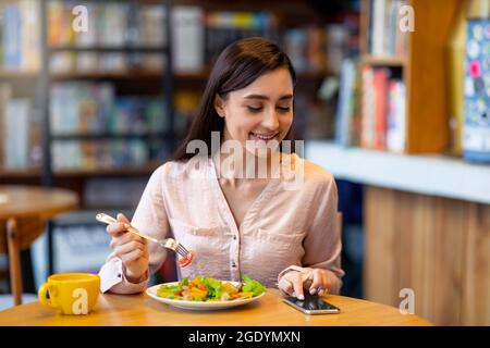 Happy young arab woman texting on smartphone while eating salad in cafe, using an application to send an sms Stock Photo