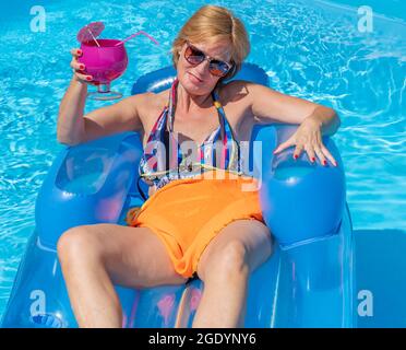 A middle-aged white woman relaxes lying on an inflatable mattress on the water of a swimming pool, with a glass of cocktail in her hand Stock Photo