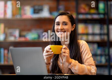 Happy arab lady enjoying coffee during work on laptop computer in city cafe, smiling and looking at camera Stock Photo