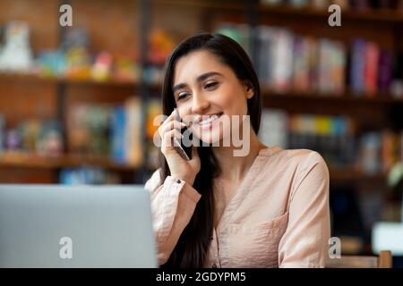 Excited latin female entrepreneur talking on smartphone while working on laptop computer in cafe, free space Stock Photo