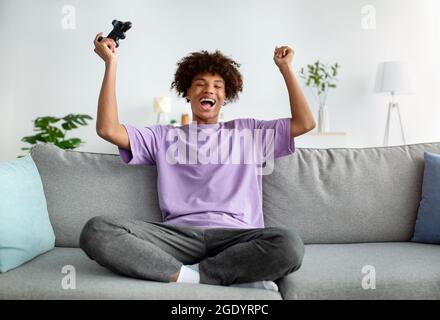 Overjoyed black teenager with joystick winning online video game, gesturing YES and screaming in excitement indoors Stock Photo