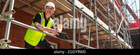 Panoramic web banner smiling male builder foreman, construction worker or architect on site wearing a white hard hat, holding a clipboard and drinking Stock Photo
