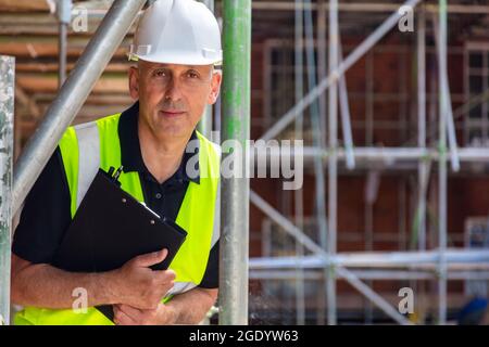 Serious looking male builder foreman, worker, contractor or architect on construction or building site holding black clipboard wearing white hard hat Stock Photo