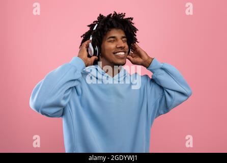 Attractive black teen guy in casual wear listening to music in headphones, closing eyes and relaxing on pink background Stock Photo