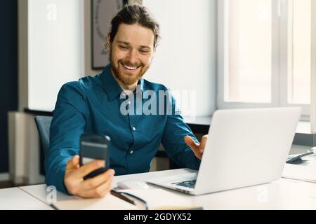 Smiling young man in casual wear looking at smartphone screen at workplace Stock Photo