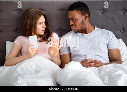 Young interracial couple having fight in bed, overusing mobile devices, having gadget addiction and family problems Stock Photo