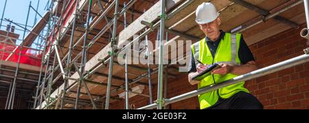 Panorama male builder foreman, construction worker, contractor or architect on building site writing on black clipboard panoramic web banner header Stock Photo