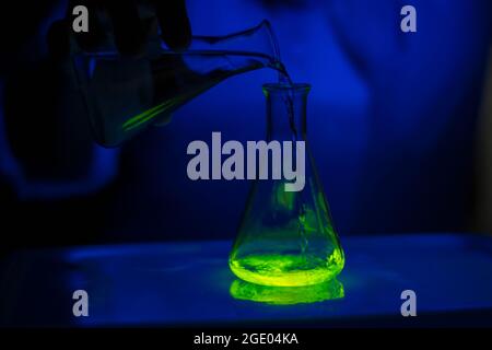A researcher experimenting with a green fluorescent compound in a glass conical flask in dark biochemistry laboratory for health care development Stock Photo