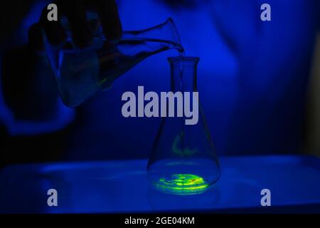 A researcher working with a green fluorescent compound in a glass conical flask in dark biochemistry laboratory for health care development Stock Photo