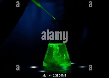 A researcher working with a green fluorescent compound in a glass conical flask in dark biochemistry laboratory for pharmaceutical development Stock Photo