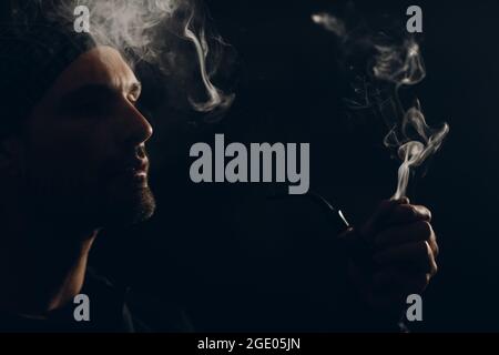 Man smokes smoking pipe against dark background. Back side lit profile portrait with smoke Stock Photo