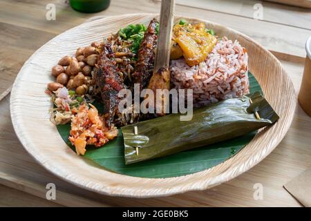 Top down view of a plate of Balinese food with fish, sate, soup, rice, sambal, greens on bamboo plate Stock Photo