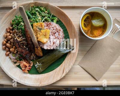 Top down view of a plate of Balinese food with fish, sate, soup, rice, sambal, greens on bamboo plate Stock Photo