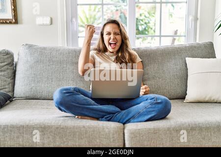 Beautiful young brunette woman sitting on the sofa using computer laptop at home angry and mad raising fist frustrated and furious while shouting with Stock Photo