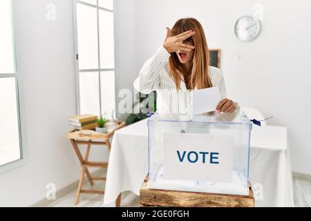 Hispanic business woman voting putting envelop in ballot box peeking in shock covering face and eyes with hand, looking through fingers with embarrass