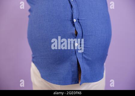 Man with overweight and tattoos holds hands on large belly on purple background Stock Photo