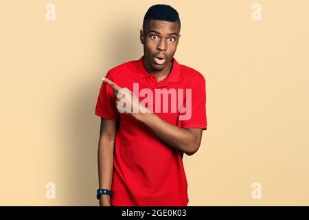 Young african american man wearing casual red t shirt surprised pointing with finger to the side, open mouth amazed expression. Stock Photo