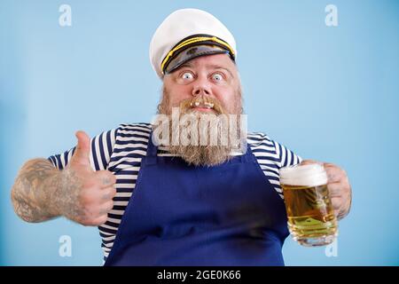 Joyful fat person in sailor suit holds beer and shows thumb up on light blue background Stock Photo