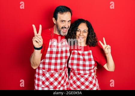 Middle age couple of hispanic woman and man wearing cook apron smiling looking to the camera showing fingers doing victory sign. number two. Stock Photo