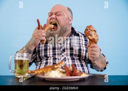 Hungry plus size man eats sausages at table with rich food and mug of beer Stock Photo