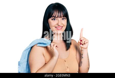 Young hispanic plus size woman wearing holding denim jacket smiling with an idea or question pointing finger with happy face, number one Stock Photo