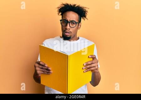 Young african american man with beard reading a book wearing glasses in shock face, looking skeptical and sarcastic, surprised with open mouth Stock Photo