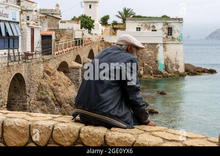 Back view of a middle-aged sitting on the rock wall, an old small lighthouse. Stock Photo