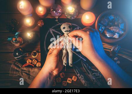 Sorceress or witch sticks needles into voodoo doll at ritual table with pentagram, burning candles and other occult objects, top view. Voodoo witchcraft, spirituality and occultism concept. Stock Photo