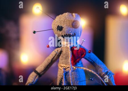 Voodoo doll studded with needles with pierced red rag heart against background of burning candles. Eerie or spooky magical esoteric ritual. Stock Photo