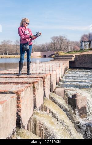 Low angle view of an adult woman standing on a brick walkway in the park holding a gimbal with phone in a sunny day. Stock Photo