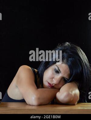 Depressed brunette woman leaning on table looking down. pensive girl. vertical, black background Stock Photo