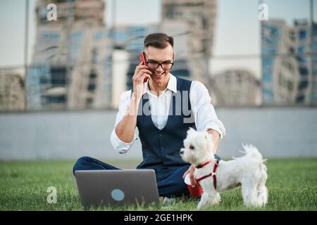man with a pet talking on a smartphone sitting on a city lawn. Stock Photo