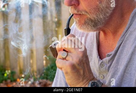 older man with facial hair smokes a pipe outdoors Stock Photo
