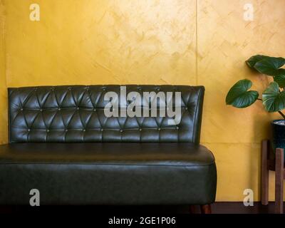 Dark green vintage leather sofa seat with pins and buttons near plant pot decoration on yellow wall background in the living room with copy space. Stock Photo