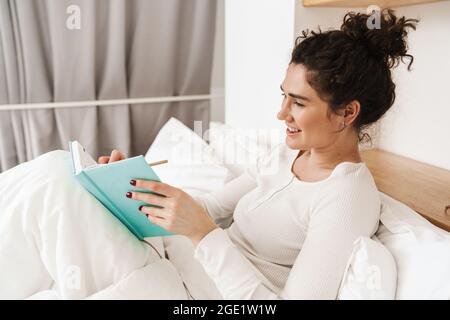 The smiling woman lying in white pajamas on the bed writing something in a diary in a bright room Stock Photo