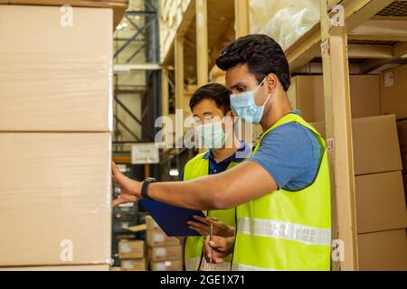 Group of young factory warehouse workers wearing a protective face mask while working in logistic industry indoor. Asian and Indian ethnic men checking item order during Coronavirus Covid 19 pandemic Stock Photo