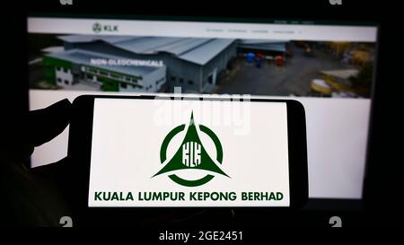 Person holding cellphone with logo of Malaysian company Kuala Lumpur Kepong Berhad (KLK) on screen in front of webpage. Focus on phone display. Stock Photo
