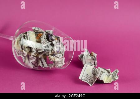 Transparent wine glass with money lying on pink background. Glass is full Crumpled hundred dollar bills. Stock Photo