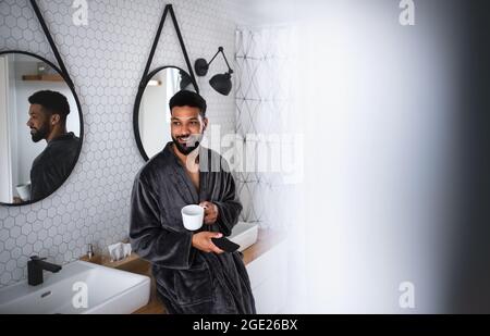 Young man with coffee and bathrobe indoors in bathroom at home, using smartphone. Stock Photo