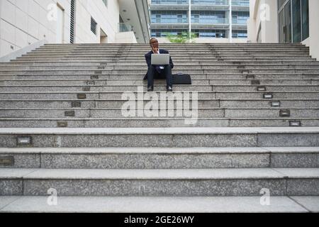 Pensive young businessman sitting on steps with opened laptop and working on important e-mail for business partner Stock Photo