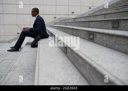 Side view of software developer in suit sitting on steps outdoors and coding on laptop Stock Photo