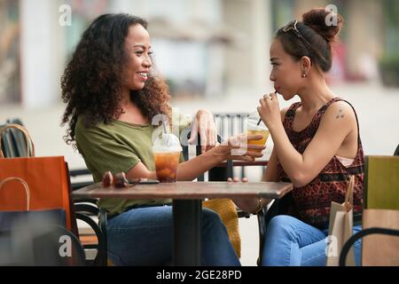 Pretty smiling young woman asking friend to taste her fruit cocktail when they are sitting at table in outdoor cafe Stock Photo