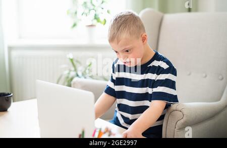 Down syndrome boy indoors at home, using laptop. Stock Photo