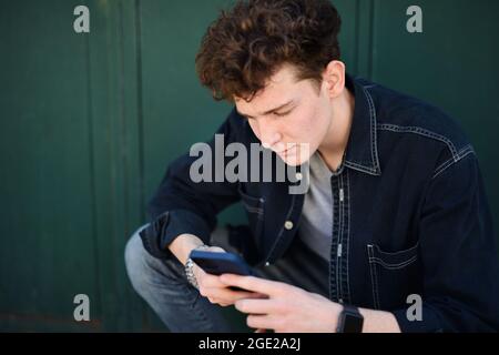 Portrait of young man against green background outdoors, using smartphone. Stock Photo