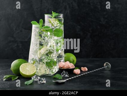 Glasses of Mojito cocktail with ice cubes mint and lime on black board with spoon and fresh limes with cane sugar. Best summer cocktail. Stock Photo