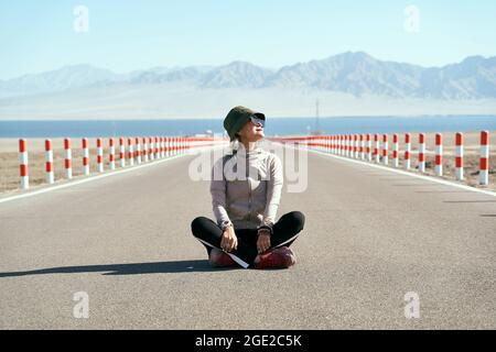 asian woman tourist sitting in the middle of an empty open road looking at view with lake and rolling mountains in background, leg crossed. Stock Photo