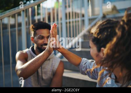 Man and woman friends resting after exercise outdoors in city, giving high five. Stock Photo