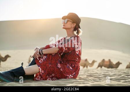 asian woman in red dress sitting in desert looking at view with caravan of camels and huge sand dune in background Stock Photo