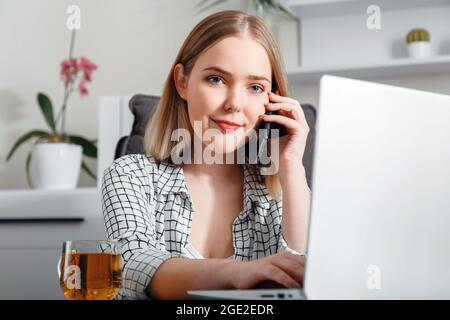 Portrait of young woman assistant or secretary talking on smartphone working using laptop at home office. Smiling woman has conversation by phone Stock Photo
