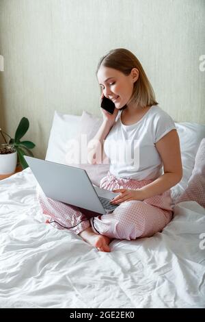 Smiling young woman in pajamas sitting in bed working use laptop smartphone in morning. Work conversation by phone during morning. Businesswoman Stock Photo